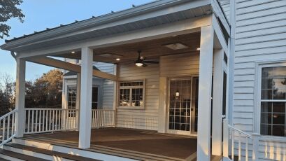 Covered porch with a wooden deck, featuring white railings, a ceiling fan, and recessed lighting. The porch is attached to a house with large windows, offering a cozy and functional outdoor space.
