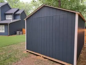 Side view of a dark blue backyard shed with white trim, standing on a grassy yard. A neighboring house is visible in the background along with a wooden fence and landscaping