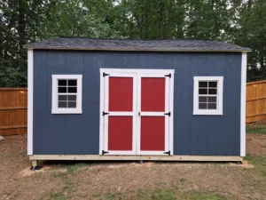 Backyard shed with dark blue exterior, red double doors, and white trim. Two windows are visible on the sides. A wooden fence surrounds the shed in the background