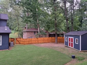 A backyard shed with blue siding and red doors, surrounded by a wooden privacy fence and tall trees. The shed is placed on a grassy area with a clear view of a neighboring house and a neatly trimmed lawn."