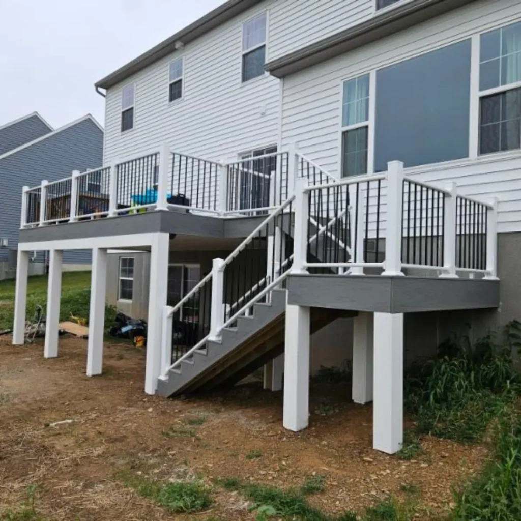 Elevated deck with white railings and black spindles, featuring a staircase leading to the ground level. The deck is supported by white columns, and the surrounding area is partially landscaped with a residential house in the background.