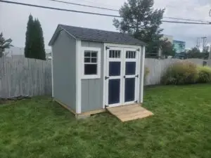 Gray outdoor shed with white-framed windows and double doors, featuring a wooden ramp. The shed is placed in a backyard with a fence in the background, offering a functional and spacious storage solution.