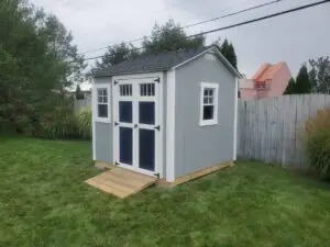 Gray outdoor shed with white-framed windows and double doors, featuring a wooden ramp. The shed is placed in a backyard with a fence in the background, offering a functional and spacious storage solution.